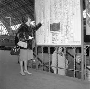 846555 Afbeelding van een vrouw die het vertrektijdenbord raadpleegt op een perron van het N.S.-station Amsterdam C.S.